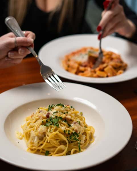 Two people enjoying creamy carbonara spaghetti and rigatoni with tomato sauce at a cozy restaurant.
