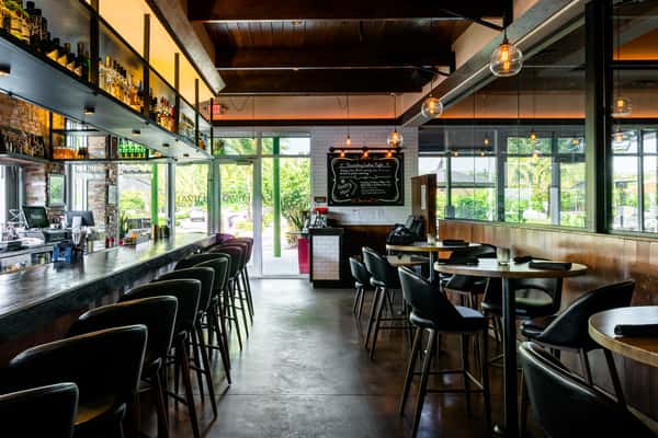 Modern bar interior with high-top tables, hanging lights, and a well-stocked bar counter near large windows with natural light.