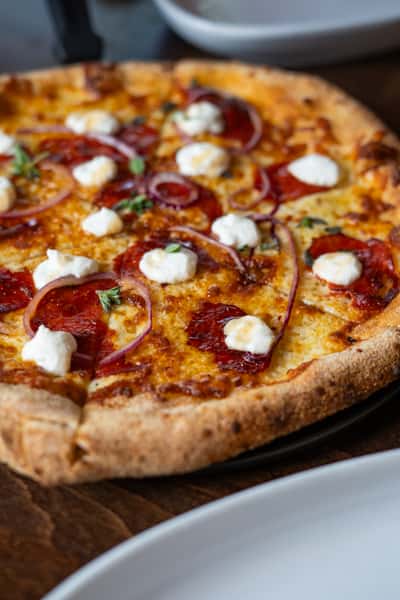 Pepperoni pizza topped with red onions, dollops of ricotta cheese, and fresh herbs on a golden-brown crust.