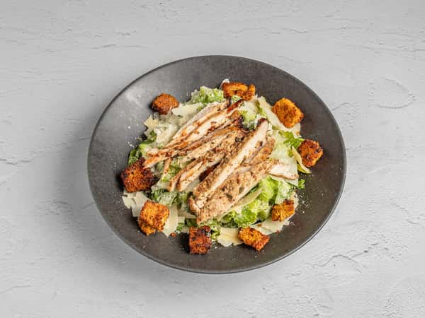This image features a fresh Caesar salad topped with grilled chicken breast, crunchy croutons, and shaved parmesan, all served in a matte black bowl for a modern presentation.