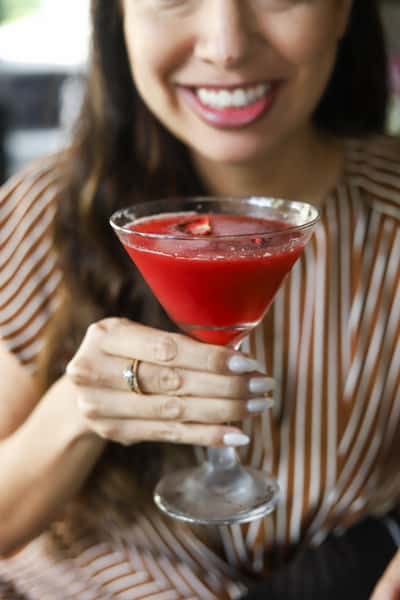 Woman smiling while holding a red strawberry cocktail in a martini glass, wearing a striped top and an engagement ring.