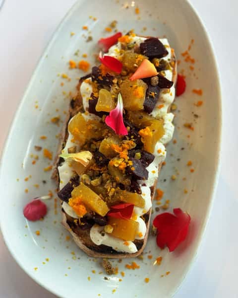 Toasted artisan bread topped with whipped ricotta, golden and red beets, crushed pistachios, orange zest, and flower petals.