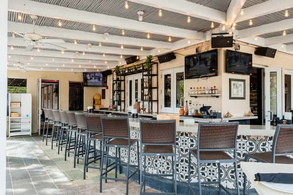 Outdoor bar with wicker-style high chairs, patterned tile bar front, string lights overhead, and mounted TVs on cream-colored walls.