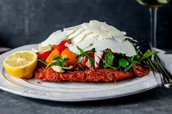 Crispy breaded chicken cutlet topped with a fresh salad of mixed greens, cherry tomatoes, and shaved Parmesan cheese, served with a lemon wedge.