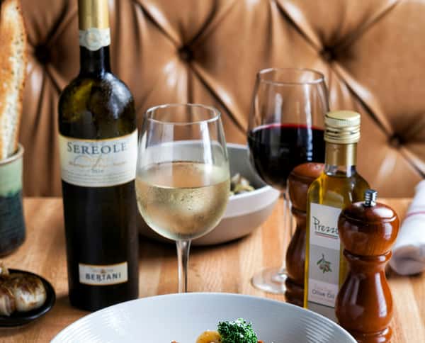 Elegant restaurant table setting featuring Sereole wine, glasses of white and red wine, Prezzo branded olive oil, and a pepper grinder, with food and bread in the background.