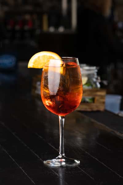 Aperol spritz cocktail with ice and an orange slice garnish served in a stemmed glass on a dark bar counter.