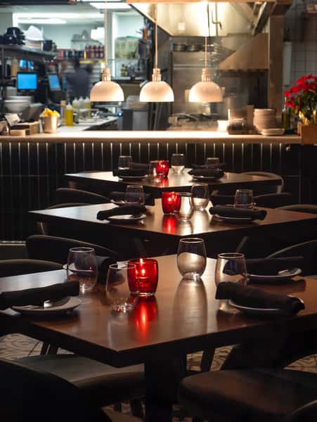 Cozy restaurant evening setting with dark wood tables, red votive candles, black napkins, and an open kitchen in the background.