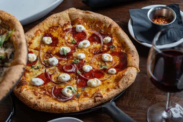 Pepperoni pizza topped with red onions, ricotta dollops, and herbs, served with red wine and crushed red pepper flakes.