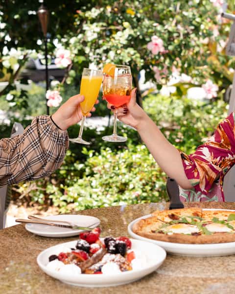 Two people clinking glasses of orange mimosa and Aperol spritz over a brunch table with waffles and pizza outdoors.