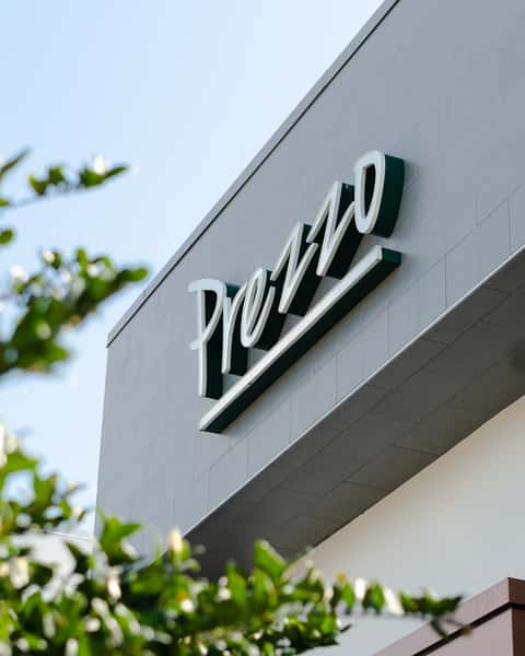 Exterior wall sign for Prezzo restaurant in bold, modern script with greenery in the foreground.