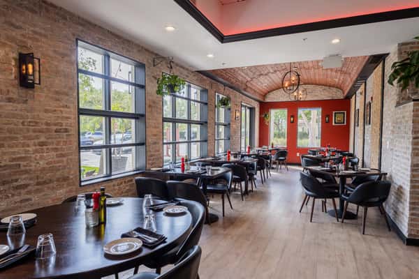 Cozy dining room with brick walls, large windows, black chairs, wood tables, and warm lighting under a vaulted wooden ceiling.