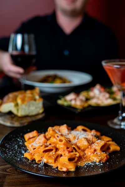 Creamy rigatoni pasta in tomato sauce topped with grated cheese, served with cocktails and bread at a cozy restaurant.