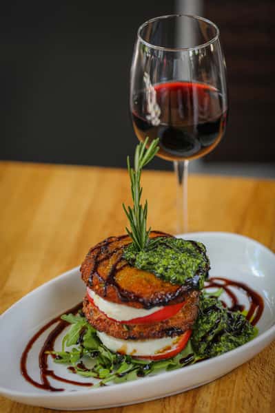 Stacked fried green tomatoes with mozzarella, tomato slices, arugula, pesto, balsamic drizzle, and a glass of red wine.
