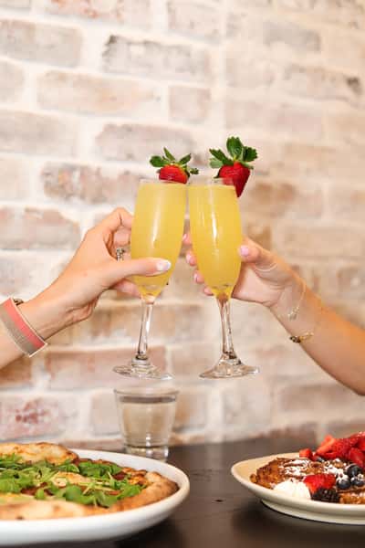 Two people toasting with mimosas garnished with strawberries over a brunch table with pizza and waffles.