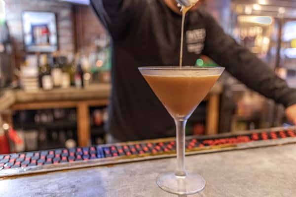 A bartender skillfully pours a cocktail into a martini glass at Gates Restaurant & Bar, with a vibrant bar scene and bottles in the background, capturing the energetic vibe.