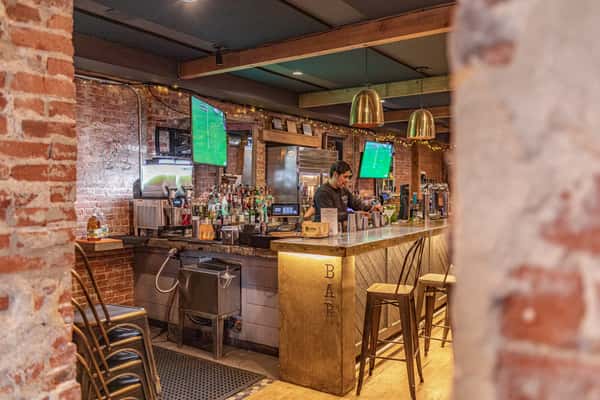 The bar area showcases a bartender preparing drinks, with shelves of liquor and multiple screens displaying sports, enhancing the lively ambiance.