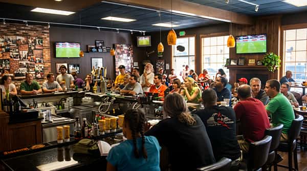 Crowded bar interior with patrons seated at a dark counter, watching televisions.