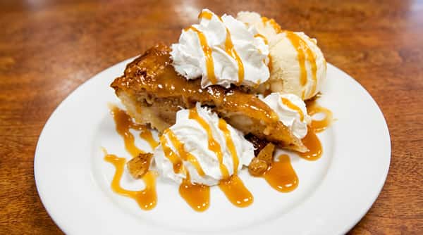 Slice of apple pie topped with whipped cream, vanilla ice cream, and caramel drizzle on a white plate.