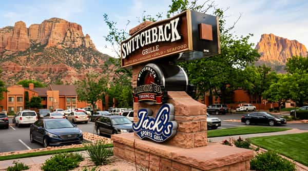 A multi-level restaurant sign featuring "Switchback Grille," "Trading Co.," and "Jack's Sports Grill" in a parking lot with mountains.