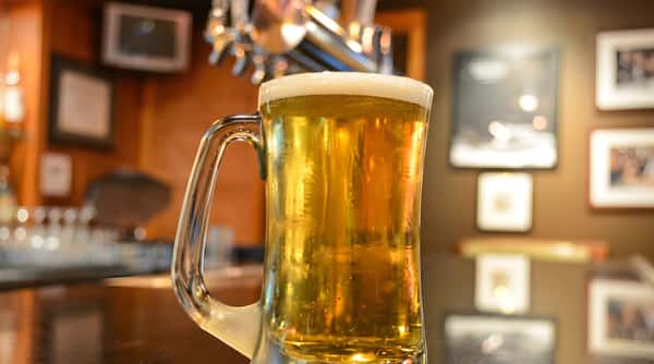 A glass mug filled with golden beer, topped with a white head of foam, in a dimly lit bar.