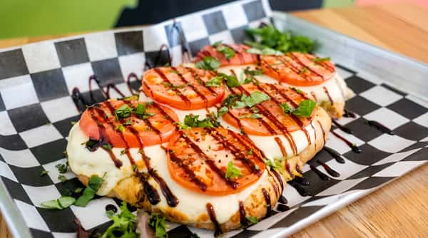Caprese bruschetta with sliced tomatoes, melted mozzarella, basil, and balsamic glaze on a checkered paper-lined tray.