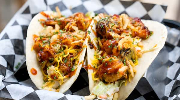 Two BBQ chicken tacos topped with shredded cheese, fried onions, and fresh parsley, in a black and white checkered basket.