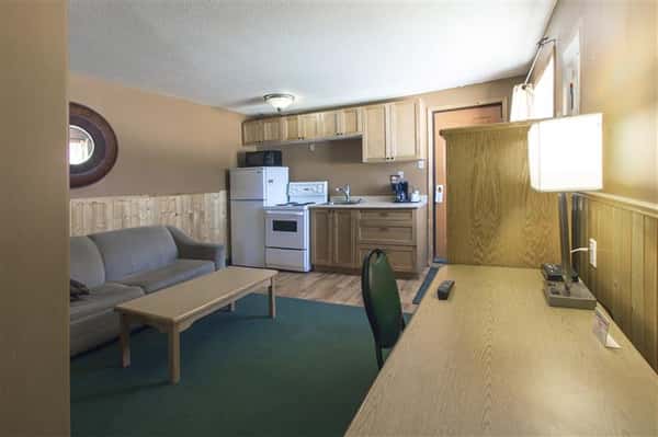 Room with couch, coffee table, desk with chair and lamp, dresser with television set, kitchen with refrigerator, oven, sink and cabinets