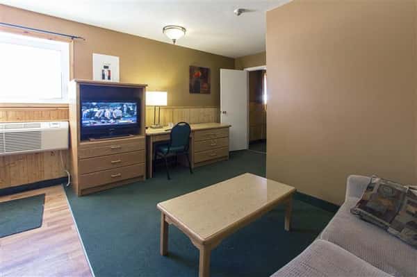 Room with couch, coffee table, dresser with television, desk with lamp and chair next to wall unit air conditioner.