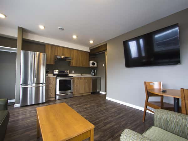 Room with wood floors, couch, chair, coffee table, wooden table with two wood chairs, television on wall and kitchen with modern appliances.