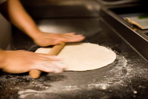 Hands rolling out pizza dough with a wooden rolling pin on a floured countertop.