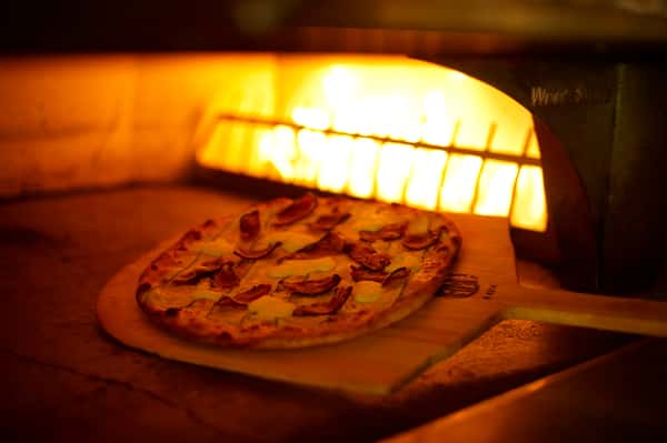 A wood-fired pizza with melted cheese and toppings being baked inside a blazing stone oven.