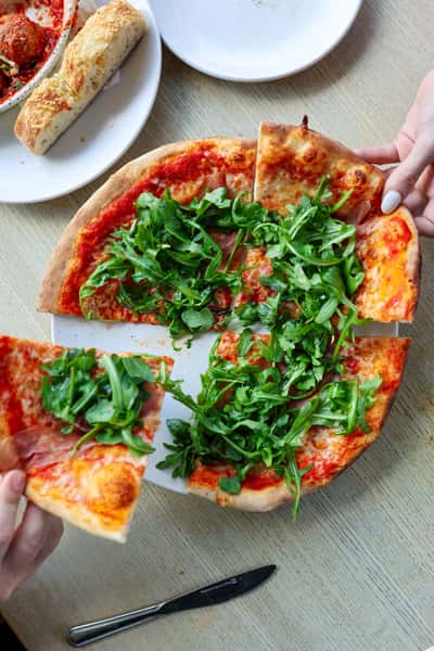Freshly baked pizza topped with arugula, with diners grabbing slices, served alongside breadsticks and meatballs in marinara.