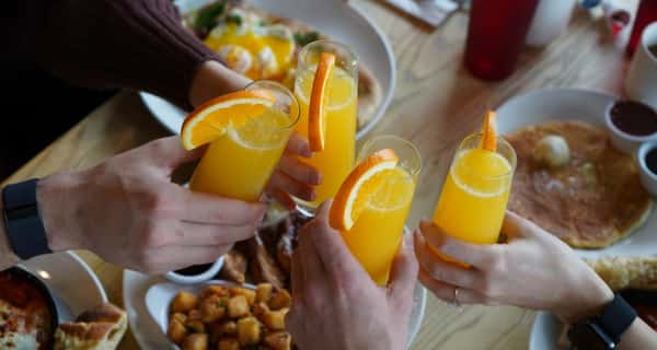 Four people clinking glasses of mimosas garnished with orange slices over a brunch spread of pancakes, eggs, and tater tots.