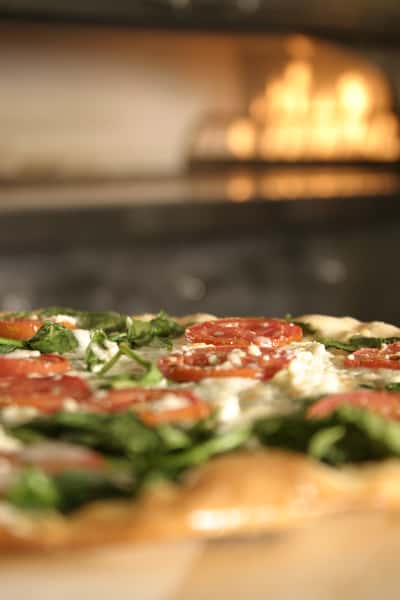 Fresh pizza topped with tomato slices, spinach, and ricotta placed near a wood-fired oven with flames glowing