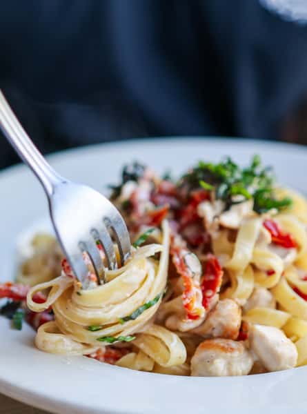 Fork twirls creamy fettuccine pasta with chicken, sun-dried tomatoes, and fresh herbs on a white plate.