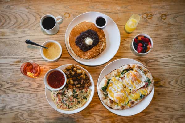 Brunch spread with pancakes, eggs benedict on flatbread, an omelet with potatoes, fresh fruit, coffee, and juice.