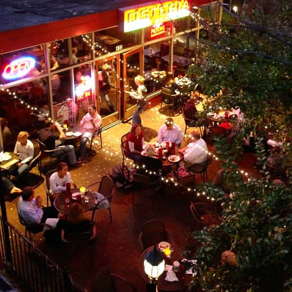 Bustling outdoor restaurant patio at night with diners enjoying meals under string lights and a glowing neon sign.