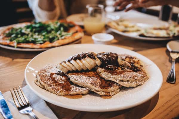 Stack of pancakes topped with sliced bananas, chocolate drizzle, and powdered sugar, served with syrup on a white plate.