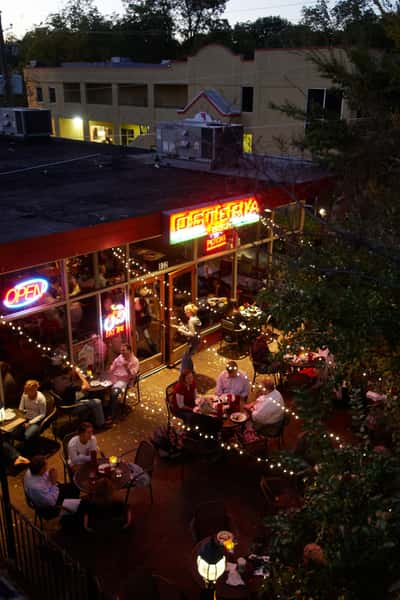 Bustling outdoor restaurant patio at night with diners enjoying meals under string lights and a glowing neon sign.