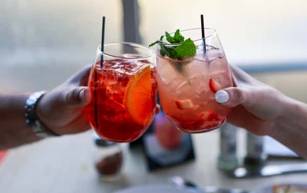 Two hands clinking glasses of colorful cocktails, one with orange slice and the other with fresh mint and strawberry pieces.