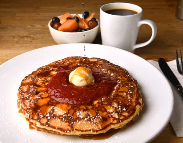 Pancakes topped with butter, strawberry sauce, and syrup are served with coffee and a bowl of fresh fruit.