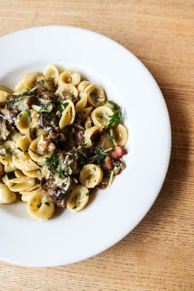 Orecchiette pasta with mushrooms, pancetta, parmesan, and parsley served on a white plate over a wooden table.