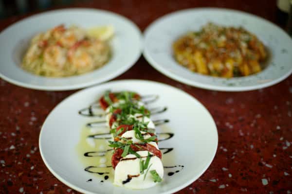 Caprese salad with fresh mozzarella, tomatoes, basil, and balsamic drizzle, served alongside pasta dishes in the background.