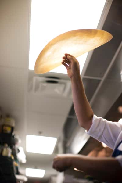 Chef stretches a thin sheet of pizza dough high in the air under bright kitchen lights, preparing it for the oven.