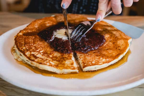 Stack of fluffy pancakes topped with melted butter, syrup, and a spread of berry compote being cut with fork and knife.