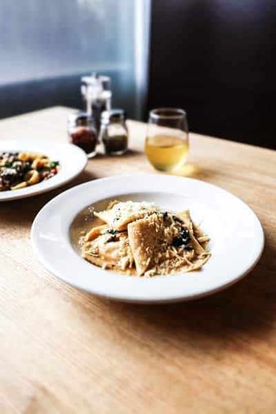 Plate of ravioli pasta topped with grated Parmesan and herbs, served with a glass of white wine on a wooden table.