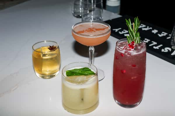 Four cocktails on a bar: a golden drink with star anise, a basil-garnished sour, a pink coupe, and a tall red drink with rosemary.