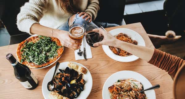 Two people toast with beer and red wine over a table of Italian dishes, including pizza, mussels, and pasta entrées.
