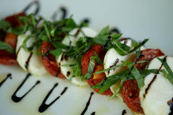 Close-up of Caprese salad with fresh mozzarella, roasted tomatoes, basil, and balsamic glaze on a white plate.