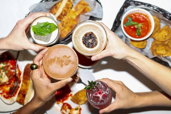 Four hands raising cocktails garnished with herbs and citrus above a table of Italian appetizers and sauces.